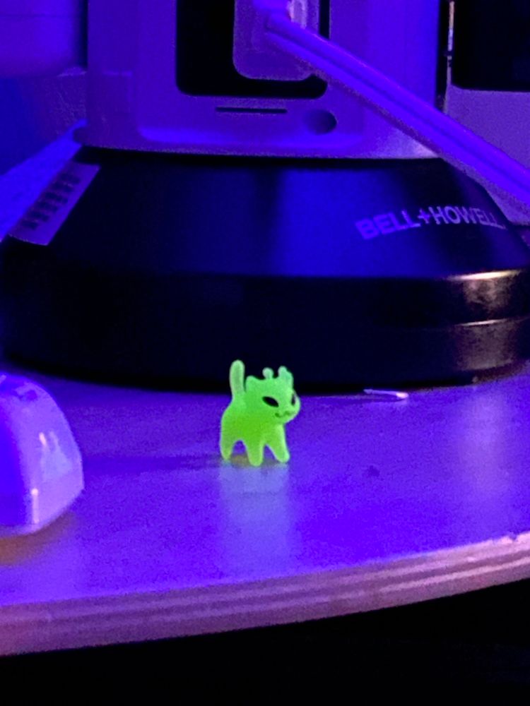 Tiny glow in the dark alien kitty figurine standing in front of a power strip thing, being very small and glowing in the dark with low purple lighting 