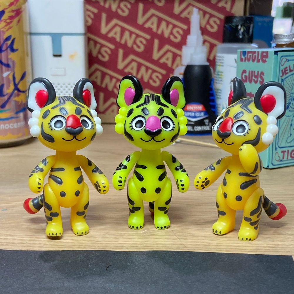 Two original-flavor Jellybean tigers (mango-yellow with charcoal stripes and red details) flank the new neon-yellow jellybean tiger with black stripes and hot pink details. 
A little secret for you who read alt-text: I finished this yellow tiger and immediately showed my wonderful partner how nice and tight the joints are with this new resin, and because it was freshly assembled it still had a little excess joint lubricant on it and almost instantly slipped out of his hands and fell on the hard floor and the arms and tail broke off and comically blasted in all directions. This is post-fix, you almost wouldn’t know it broke 😅
I have no idea why I don’t have soft rugs covering every inch of my office floor at this point, I’ve lost many a guy to my own butterfingers and this hard, unforgiving laminate flooring 🫠