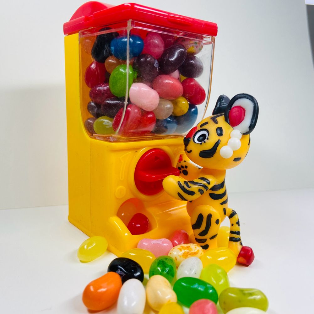 A small tiger figurine standing at a toy candy dispenser full of jellybeans, with beans overflowing out of the receptacle thingy  