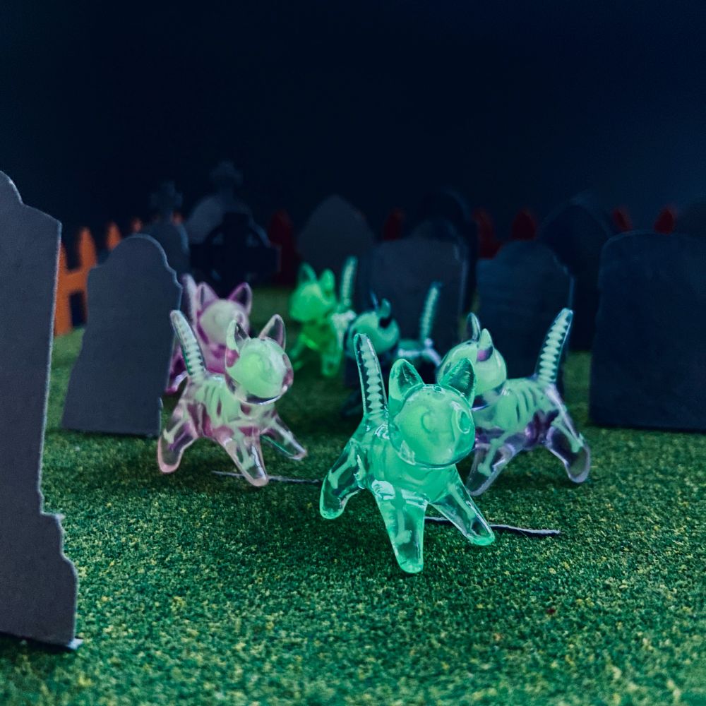 6 small translucent cat figurines with glow in the dark skeletons visible inside them,  standing among some paper headstones in a miniature graveyard scene