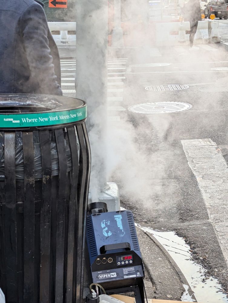 A theatrical fog machine on a city street next to a trash can. It is spewing a thick grey haze.