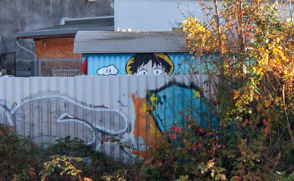 Wellblechzaun mit Graffiti, dahinter ein bemalter Bauwagen, von dem ein Teil (ein junger Mangadude, aus Dragon Ball? hab keine Ahnung) sichtbar ist.