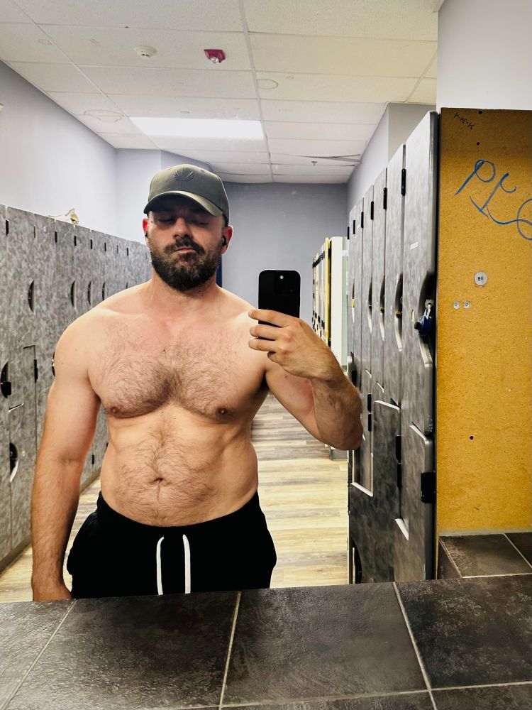 Bearded guy in green hat and black shorts, taking photo in empty locker room mirror.