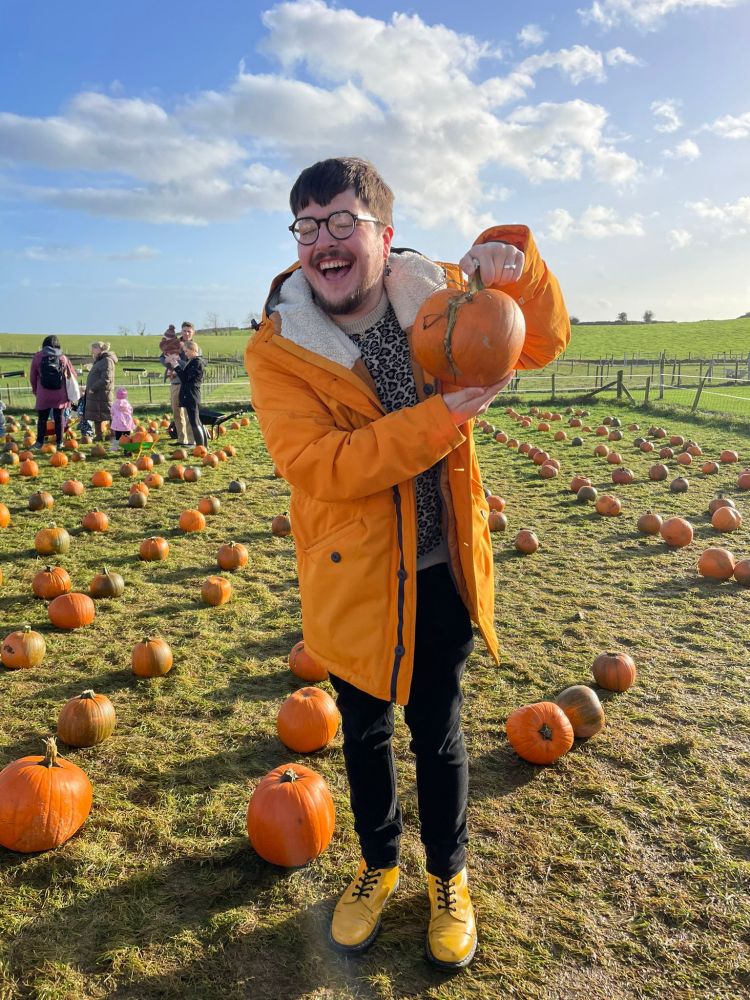 A man in his thirties is standing in a pumpkin patch wearing glasses, a bright yellow coat, bright yellow boots. He is holding a pumpkin that resembles a human rear and is laughing.