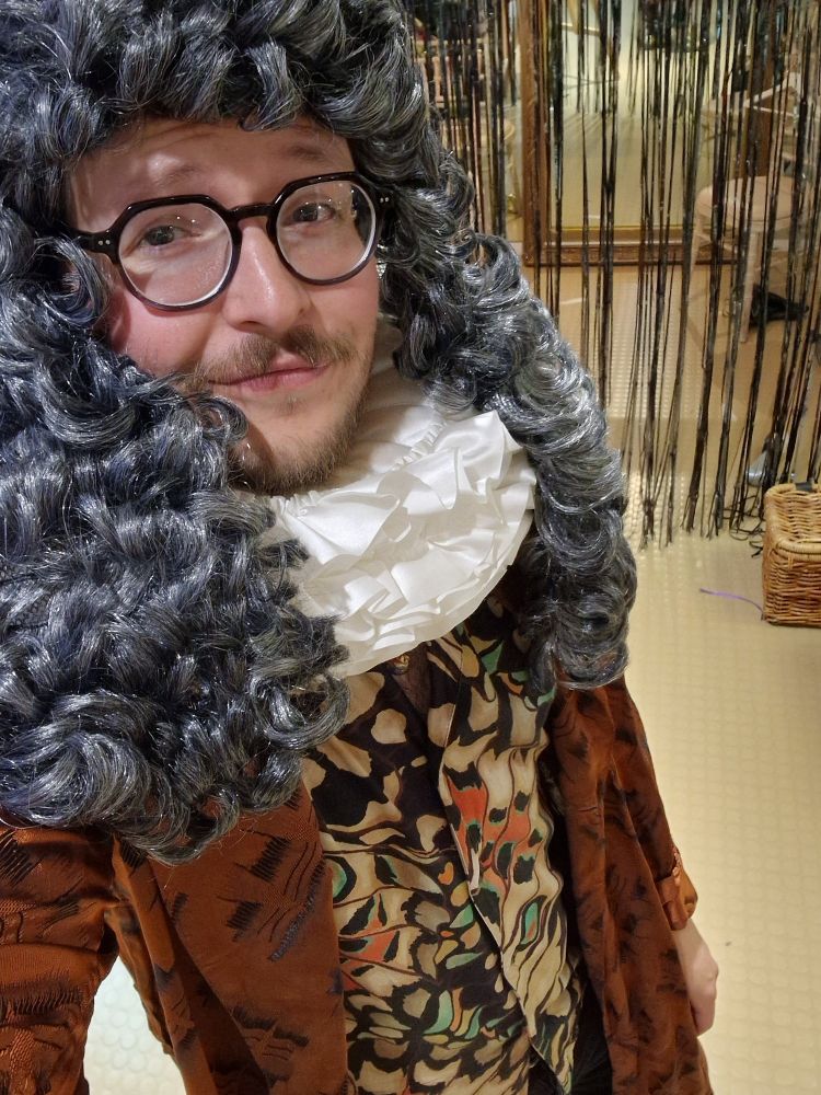 A man in his thirties with a beard, moustache and glasses is wearing a period fancy dress costume including a brown coat, white neck ruff and long curly silver wig.