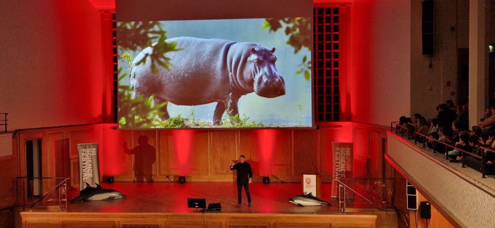Steve Backshall, a man wearing black jeans and a black top, presents from the centre of the stage. He is backed by red lighting and the projector screen shows a photograph of a hippo.