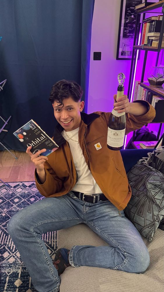 Gabe on the sofa holding a bottle of wine and the book Orbital by Samantha Harvey 
