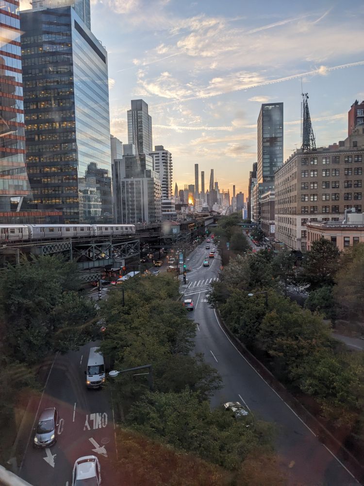 View from the train in Astoria, NYC. Tree-lined roads with the elevated train tracks and cityscape around them at sunset. 