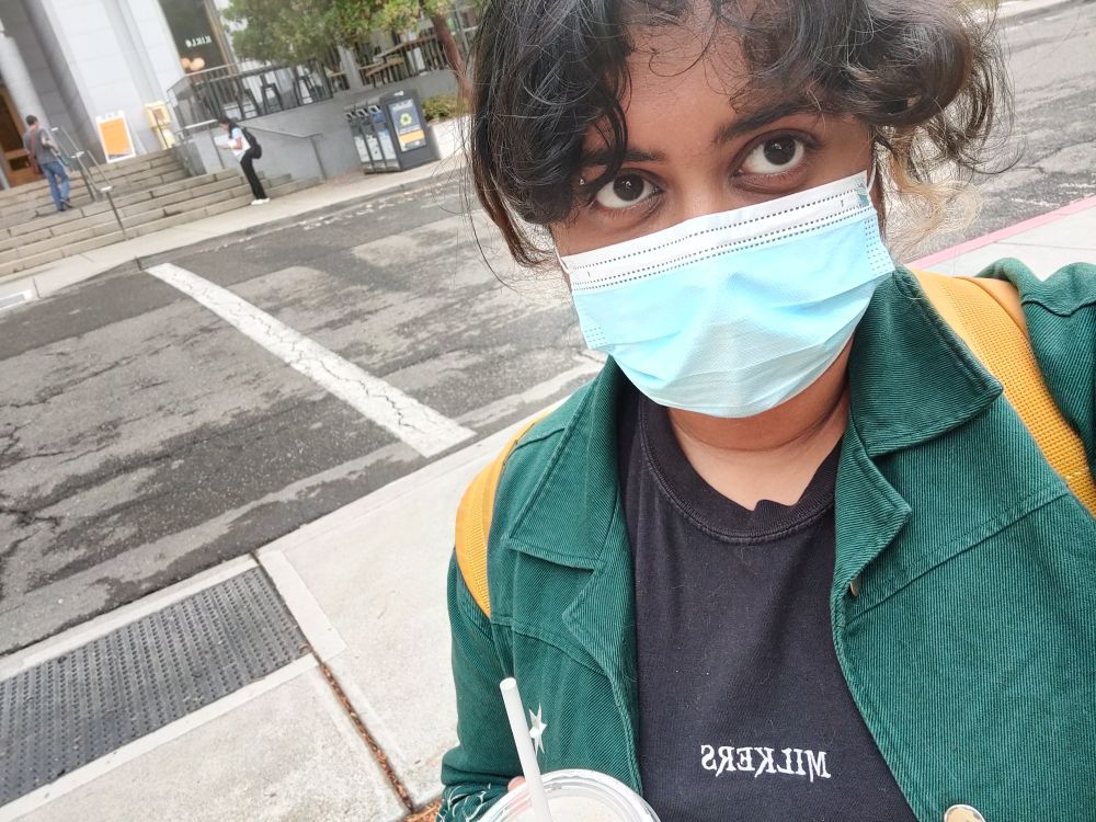 Selfie of me at a crosswalk with a blue surgical mask, a black shirt that has the word milkers embroidered on, a green jacket, and yellow backpack straps over my shoulders.