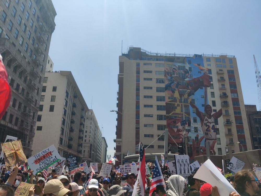 Large crowd of people with various signs and flags protesting. Mural of Kobe Bryant in the background.