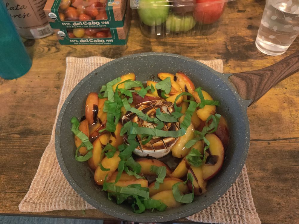 Yellow peach slices with a wheel of scored brie cheese in the middle. There is a brown balsamic glaze drizzled on and fresh green basil leaves sprinkled on top of everything. The dish rests in a blue grey pan. The pan has a brown wooden handle. The pan sits atop a tan kitchen towel that is placed on a wooden table. There are some packaged tomatoes in the background. 