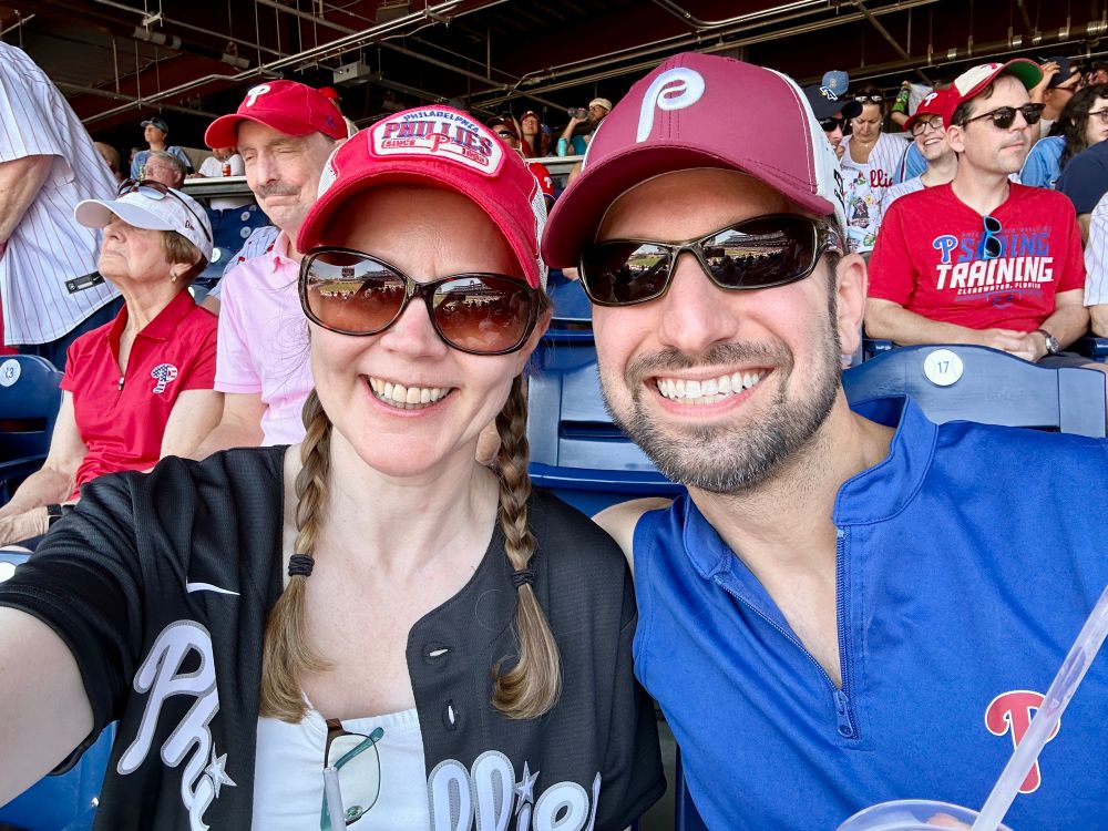 A man and woman smiling wearing Philadelphia Phillies gear in the stands for a game.