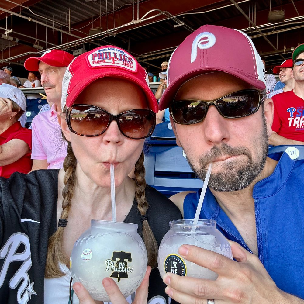 A man and woman sipping frozen margaritas wearing Philadelphia Phillies gear in the stands for a game.