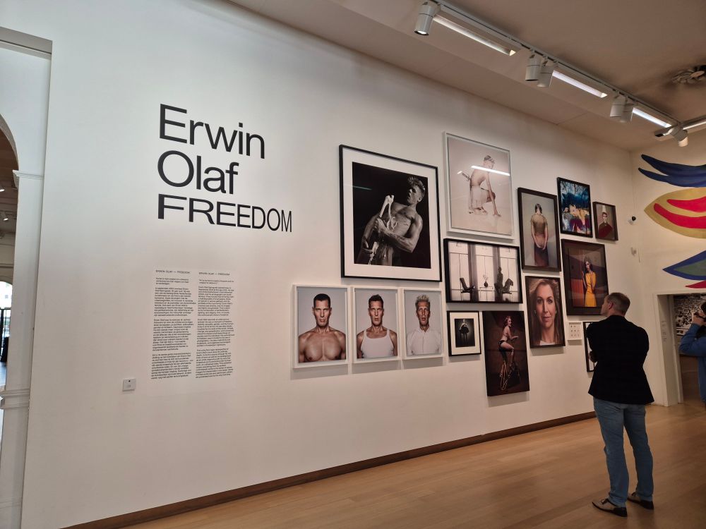 Grote witte wand in het Stedelijk Museum met in grote zwarte letters Erwin Olaf Freedom en daarnaast een verzameling van verschillende kleuren en zwart wit foto's uit het oeuvre van kunstenaar Erwin Olaf.
