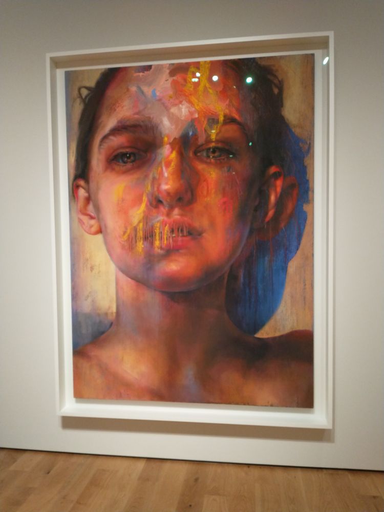 A portrait by Jenny Saville, in oils on linen, over drawn with pastels. It shows a portrait of a young woman, her lips seem almost be stitched closed using bright pastel wax. Her eyes, like almost all of Savilles' portraits, are deep and haunted.