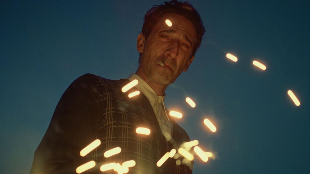 Scene from the 2024 film The Brutalist. Lead actor Adrian Brody—a white man in his late 40s or early 50s with short dark hair, wearing a dark plaid sweater over a white button-up collared shirt—set against a dark blue sky at dusk, looks down at the camera, cigarette in his mouth, trademark squint on his face as embers swirl in front of him, indicating a fire just out of frame.