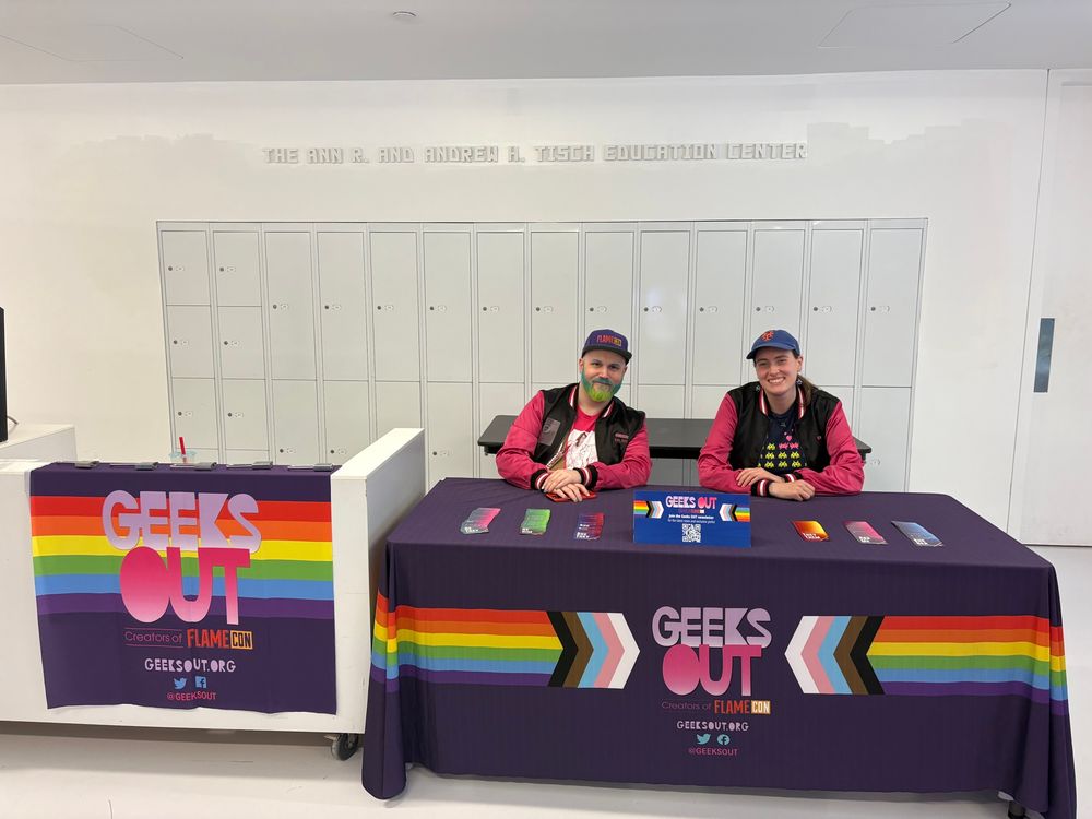 Members of Geeks OUT sitting at a table decorated with a Progress Pride rainbow Geeks OUT tablecloth. 