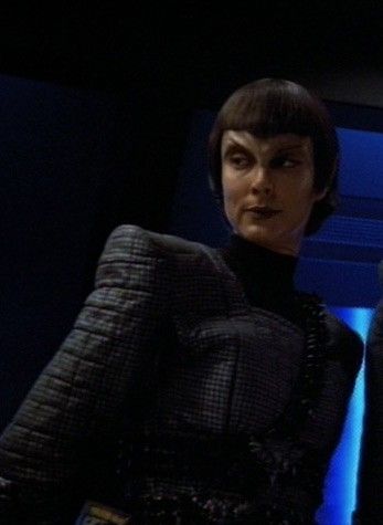 Commander Nevala, Romulan, in a dark, quilted uniform, hands behind her back, with blue lighting behind her left arm, looking judgemental