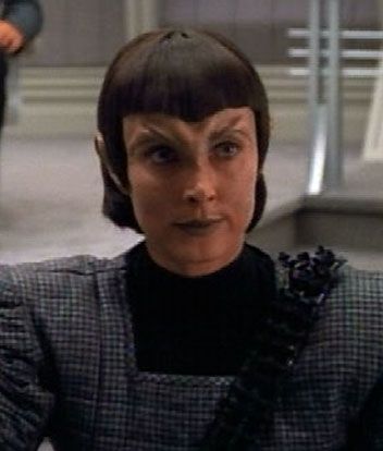 Nevala, dressed in a dark grey checked uniform with a square black collar, and spiked black sash. Her lips are painted dark, hair in a military crap 