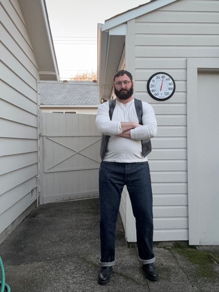 Facing towards the camera, posing outside in a leather vest, jeans, a long-sleeve white shirt, and leather boots. Arms are crossed.