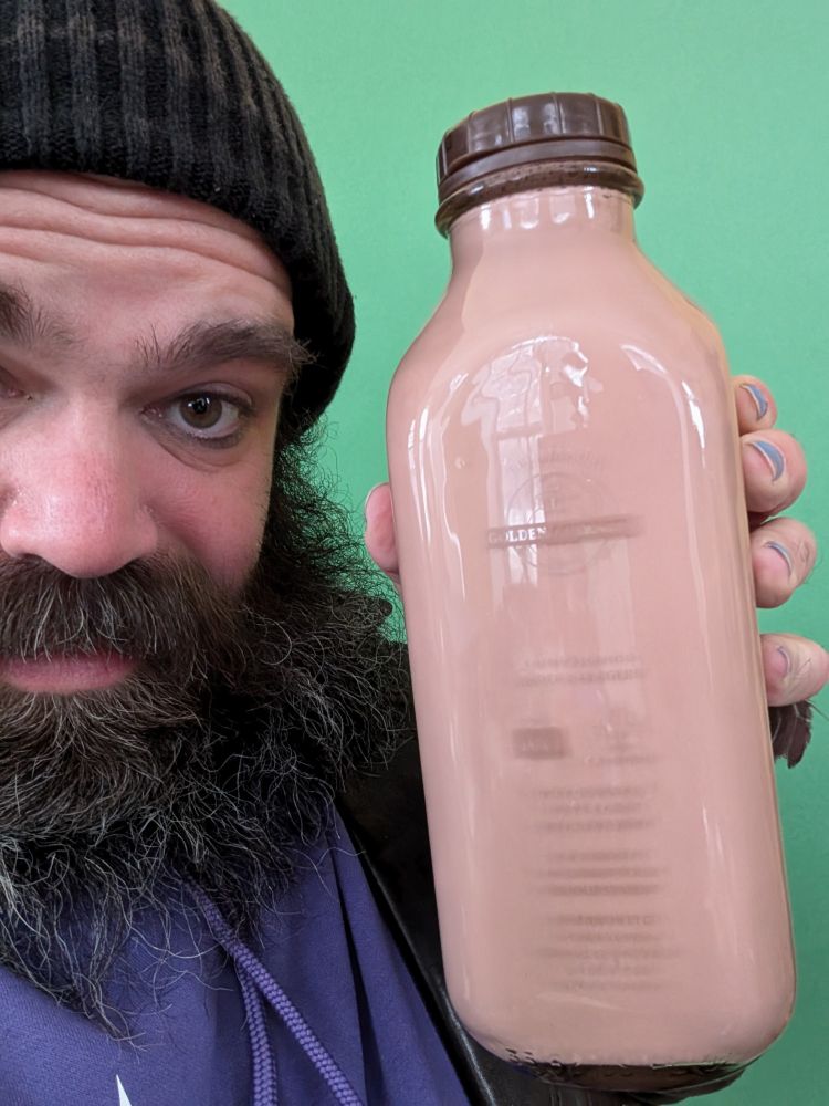 A bearded man, half out of frame, holding a glass bottle of milk in front of a green screen.