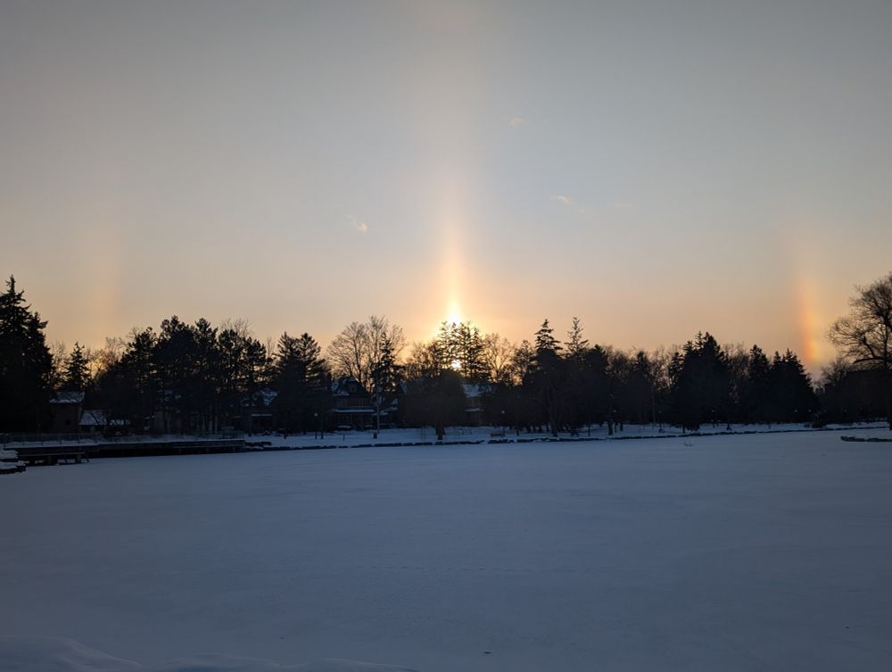 Winter halo in Victoria park downtown Kitchener. The sun is in the center and a rainbow arc is on either side. the foreground is a frozen lake.