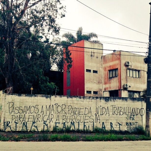 O muro de uma fábrica e nele está pixado: “PASSAMOS A MAIOR PARTE DA VIDA TRABALHANDO PARA ENRICAR A BURGUESIA”