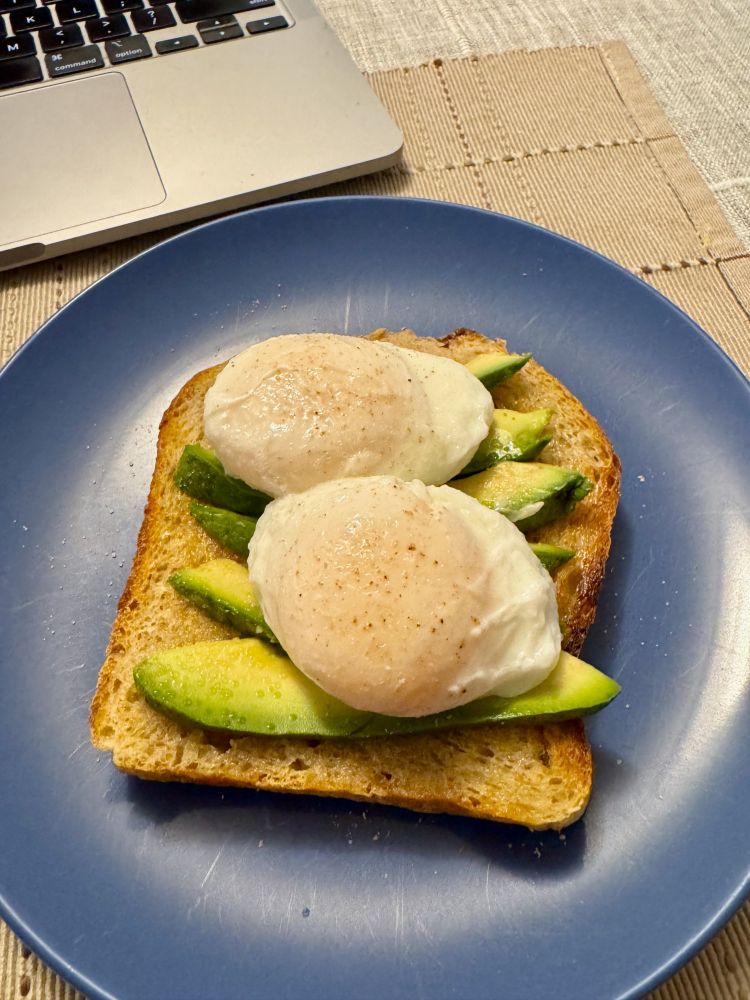 A picture of two poached eggs on top of sliced avocado, on a piece of whole wheat bread. The plate they are sitting on is blue. In the upper left corner, there is the edge of a laptop.