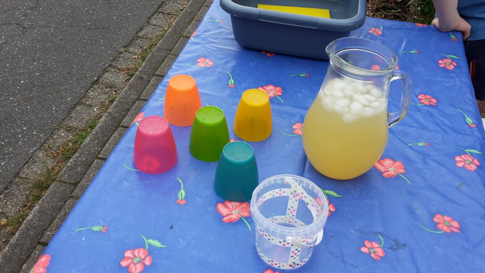 Auf einem Tisch mit einer blauen Tischdecke mit Blümchen stehen 5 bunte Trinkbecher und 1 Krug mit selbst gemachter Limonade mit Eiswürfeln. Der Tisch steht am Straßenrand. 