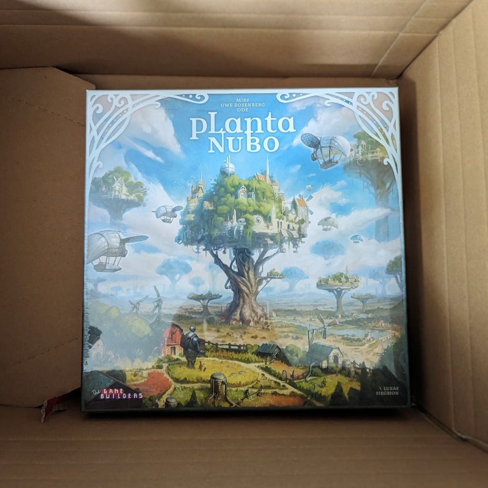 Die Schachtel des Spiels Planta Nubo. Im Zentrum steht ein großer, in die Wolken ragender Baum. Seine Krone ist mit Häusern bebaut und Luftschiffe fliegen durch nahe Wolken. Im Hintergrund sind schemenhaft weitere riesige Bäume zu sehen, die ähnlich bebaut sind. 
Im Vordergrund sind landwirtschaftliche Flächen zu sehen und eine Figur, die am ehesten an einen Androiden erinnert, der, in landwirtschaftlicher Betätigung, in Richtung des großen Baumes schaut.