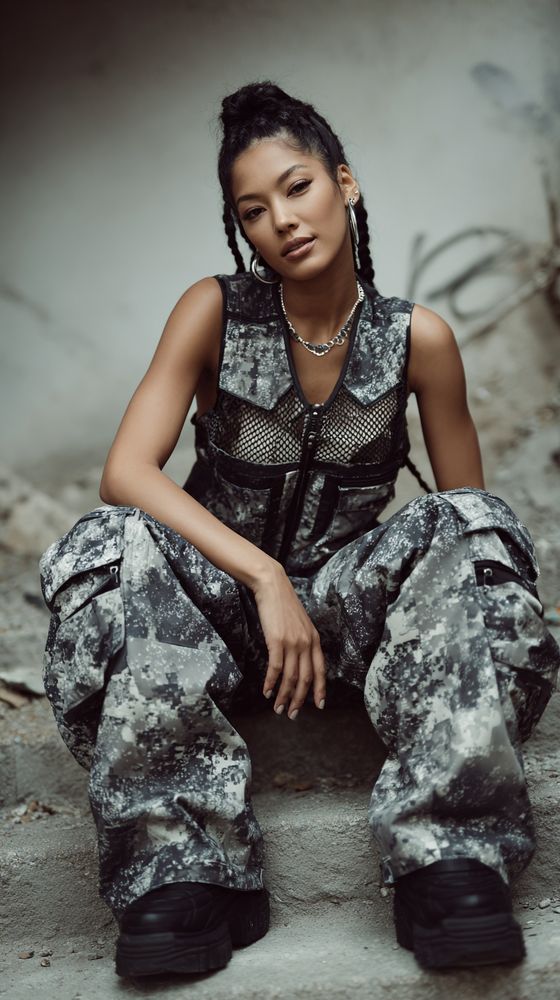 fashion model in grayscale digital camo pants and mesh panel tactical vest, seated on weathered concrete steps with a soft-focus graffitied wall behind her, braided high bun, silver jewelry, moody lighting, 85mm lens, f/2.0 for shallow depth of field, editorial streetwear aesthetic
