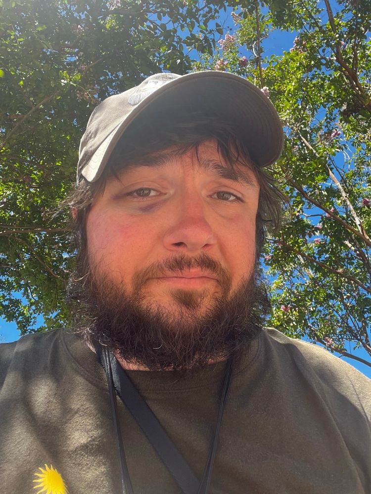 a selfie of me looking at the camera, outside under the diffused shade of the tree with sunlight also glinting in through the branches and leaves. 