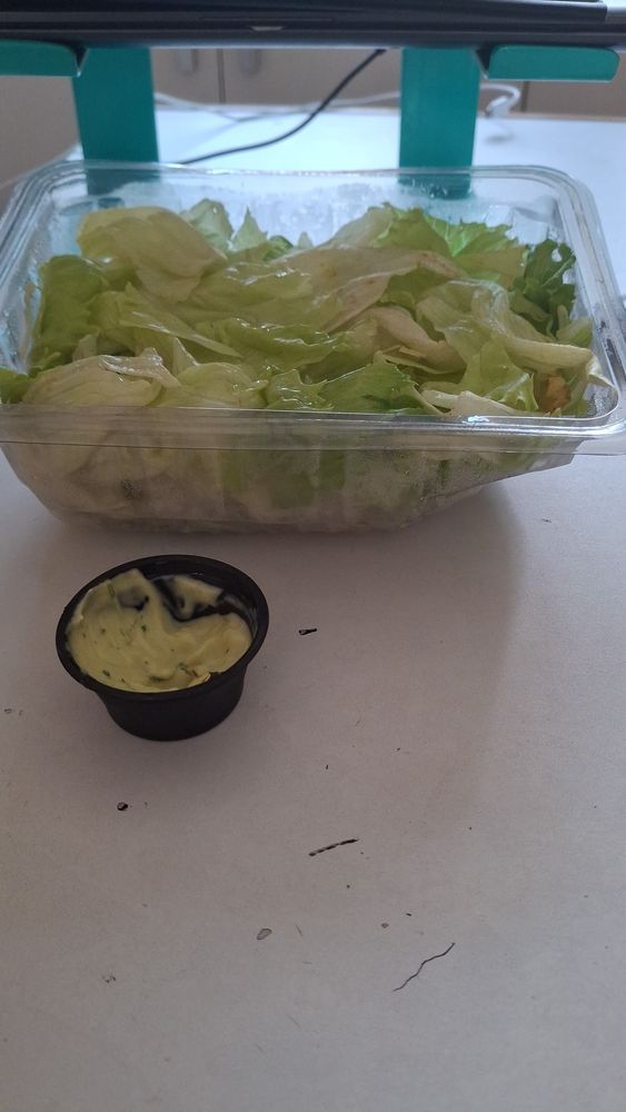On a white surface, an open plastic container of iceberg lettuce and a smaller, black container of maionese verde (kind of mayonnaise with herbs, common in brazil)