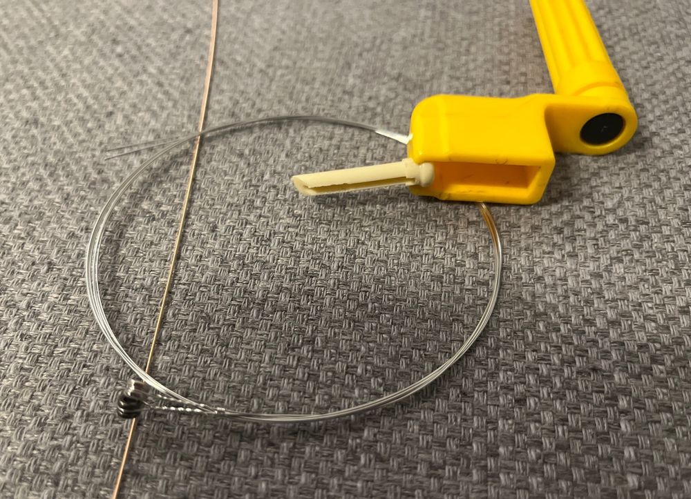 Man sieht drei neue Gitarrensaiten, zwei davon noch aufgewickelt, Zusätzlich eine Saitenkurbel und einen Bridge-Pin.