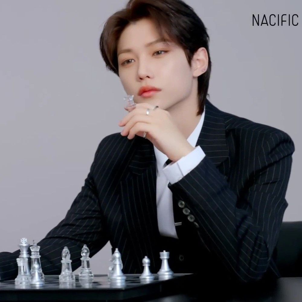 felix in a black grey-striped suit holding  a chess piece, he is seated and there's a black table in front of him with a chess board