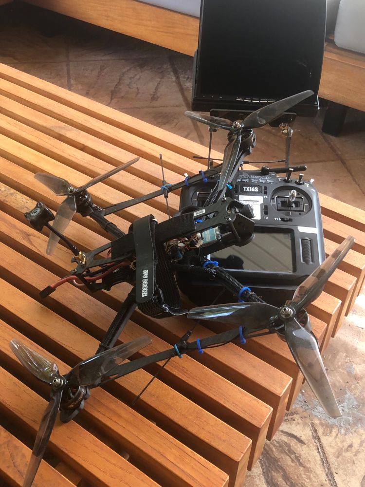 A custom-built quadcopter drone with four large propellers is resting on a wooden slatted bench. The drone is secured to a black TX16S remote controller using a black "FPV DONE" battery strap and several blue zip ties. The controller features dual joysticks, various switches, and a built-in screen, with an attached FPV monitor mounted above it. The setting appears to be an indoor or semi-outdoor lounge area with stone tile flooring and wooden furniture.