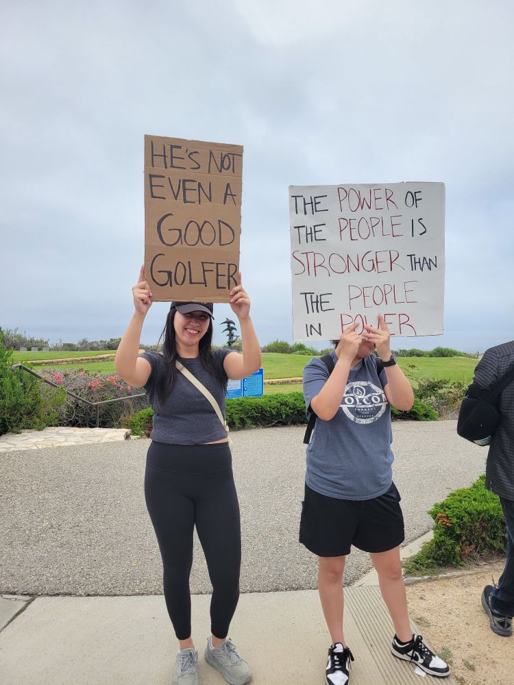 Two women holding signs. 
One says "He's not even a good golfer" and the other sign says 
"The power of the people is stronger than the people in power"