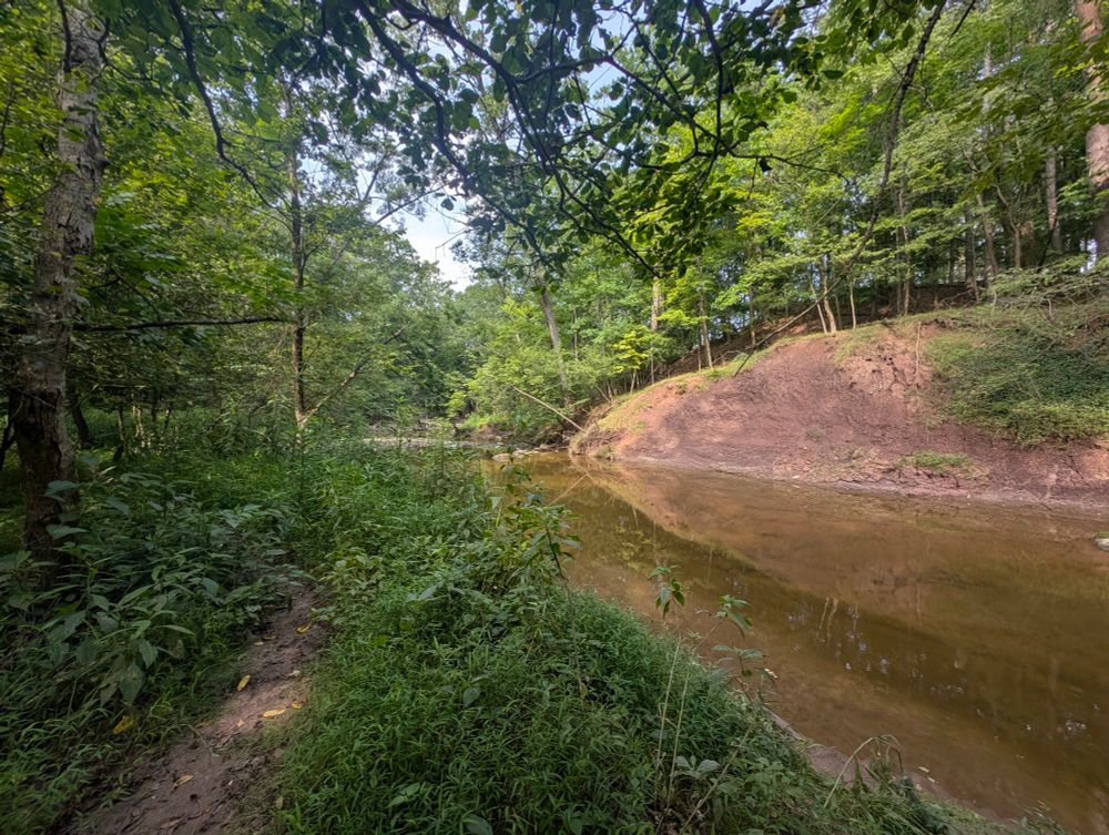 A path next to a creek, with a cliff of purple-red clay across the water