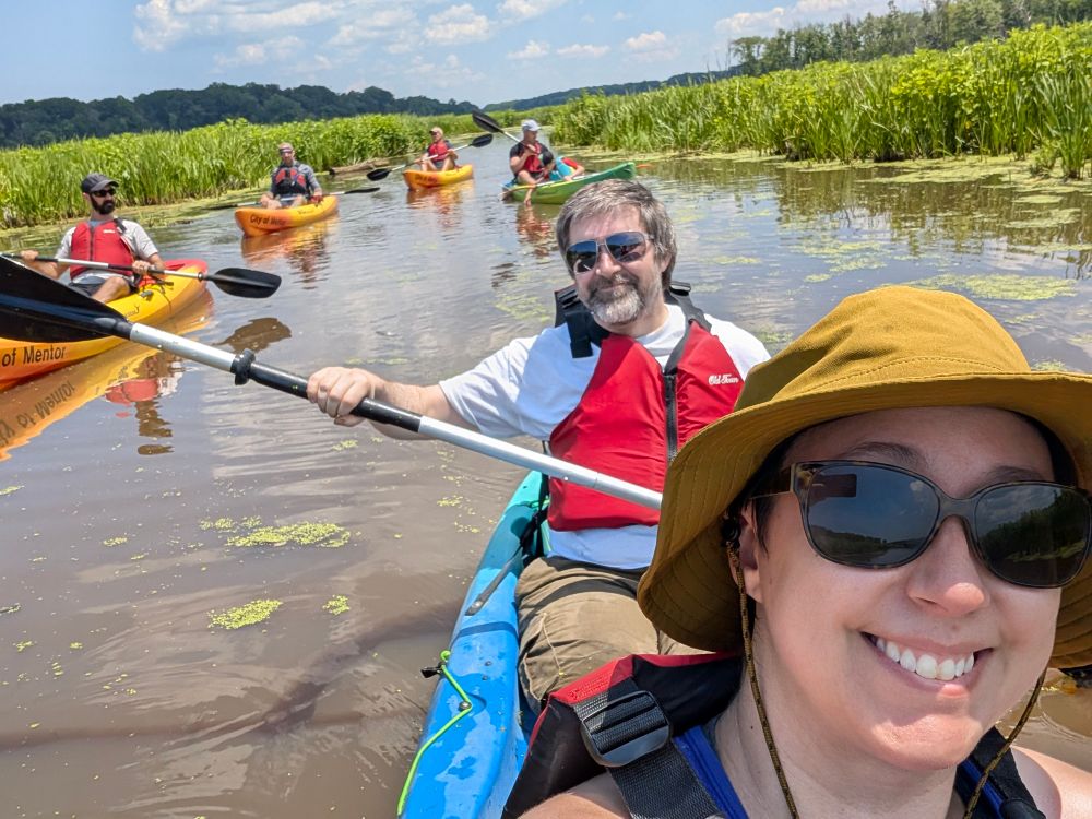 A selfie of a man and woman kayaking in a marsh with four more kayaks in the background 