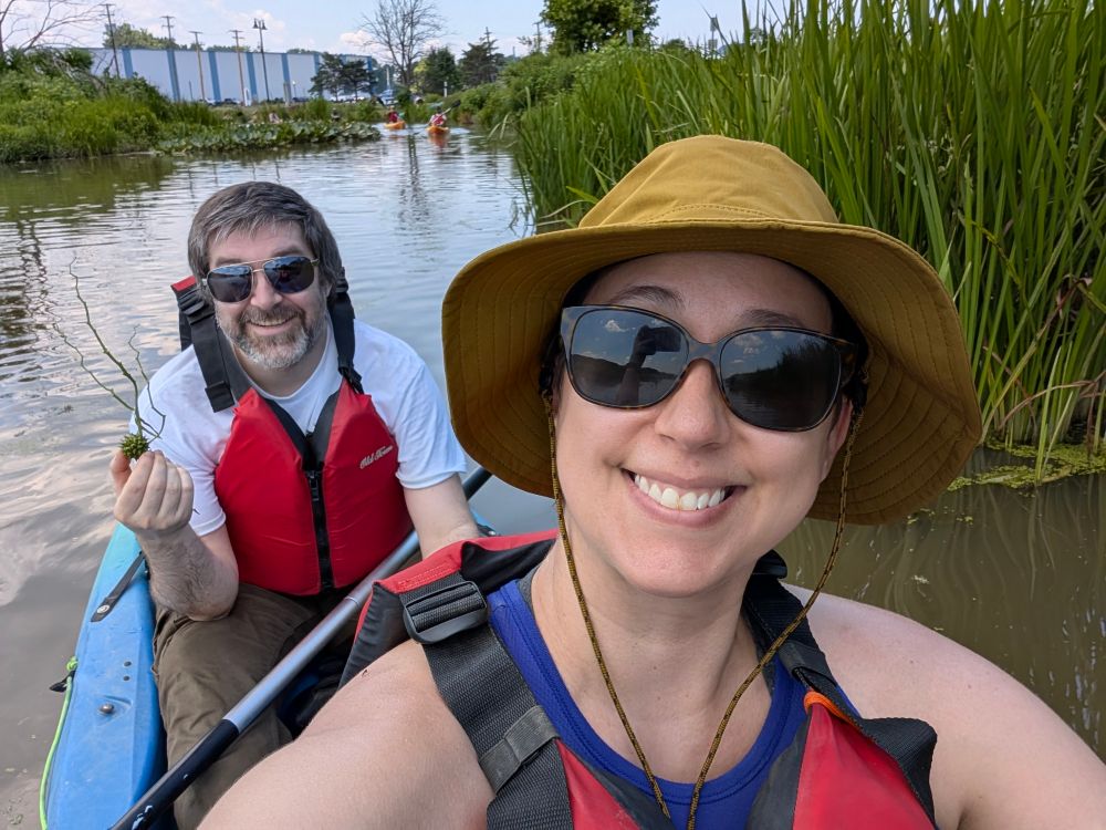 A selfie of a man and a woman kayaking. The man, in the back of the kayak, is excitedly showing off the plant pod he just discovered.