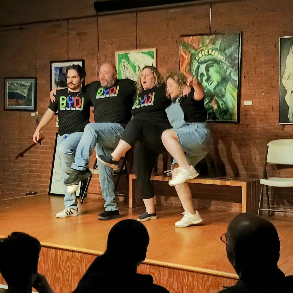 A classic kickline moment! Four cast members link arms and lift a leg mid-kick as the audience watches and laughs along.