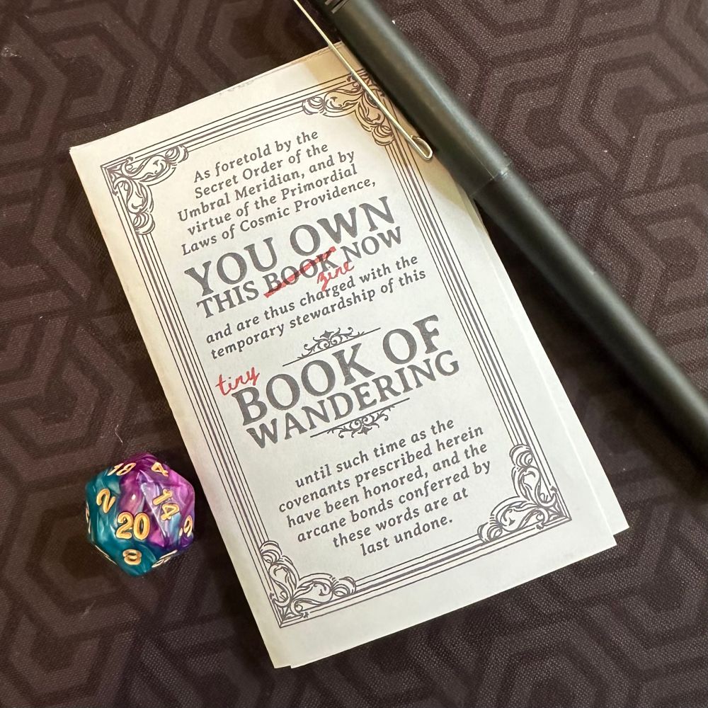 A folded zone-style Tiny Book of Wandering with a D20 on one side and a pen on the other. 