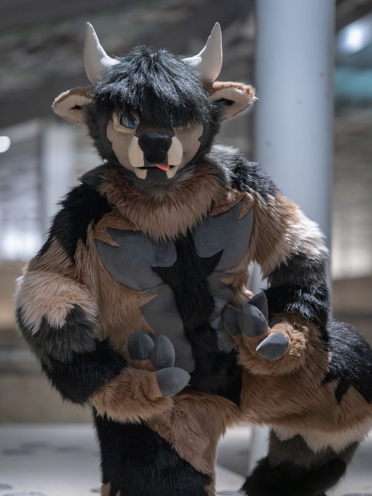 An adorkable Earth colored bull fursuiter, trying his best to flex. His tongue is sticking out slightly, and his hair is covering his eyes