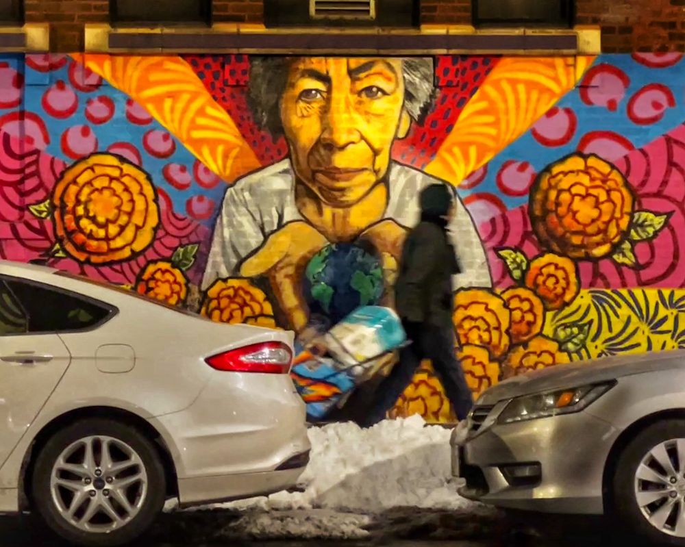 Large mural in Albany Park, Chicago, depicting an elderly person gently holding the Earth; warm orange and red motifs and oversized flowers surround the figure, conveying themes of care, wisdom, and environmental stewardship
