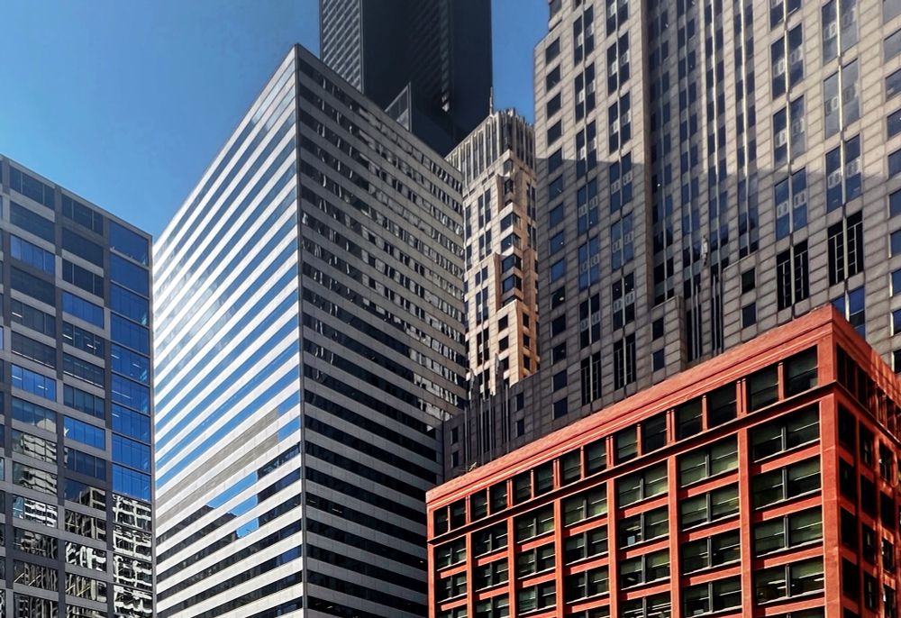 A sampler of Chicago Loop skyscrapers: modern white-and-gray striped office tower, a red-framed glass building, and textured masonry facades in the foreground, with the dark, bundled-tube “Sears” Tower standing tall in the background beneath a clear blue sky