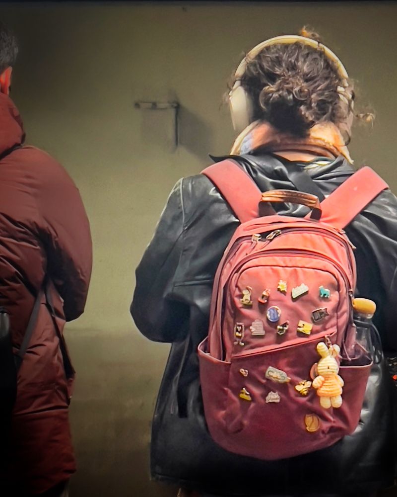 Two people stand on a CTA platform seen from behind. The right figure wears large headphones, a dark leather jacket, and a red backpack covered in colorful pins and a plush keychain.
