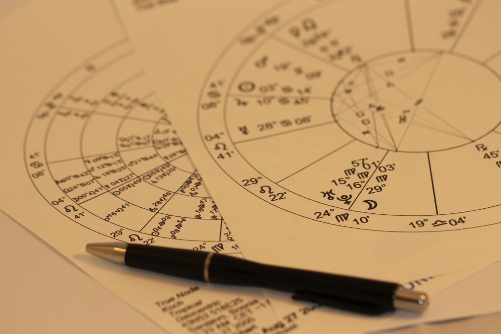 A close-up photo of printed astrology birth charts with planetary symbols and house divisions, partially overlapped, with a black pen resting on top.