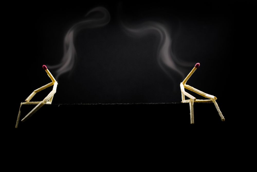 Two burnt-out matchstick figures sit facing away from each other on a black background, with smoke rising from their heads, symbolizing emotional exhaustion and estrangement.