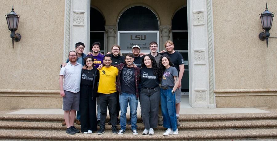 Twelve smiling graduate students standing on the steps in front of the LSU Museum of Natural Sciences. Back (L to R): Jerry Su, Gustavo Martins, Samantha Rutledge, Quinn McCallum, Ryan Klutts, David Vander Pluym. Front (L to R): Eamon Corbett, Juliana Damasceno, Diego Cueva, Luca Micheli, Amanda Harvey, Sara Velásquez