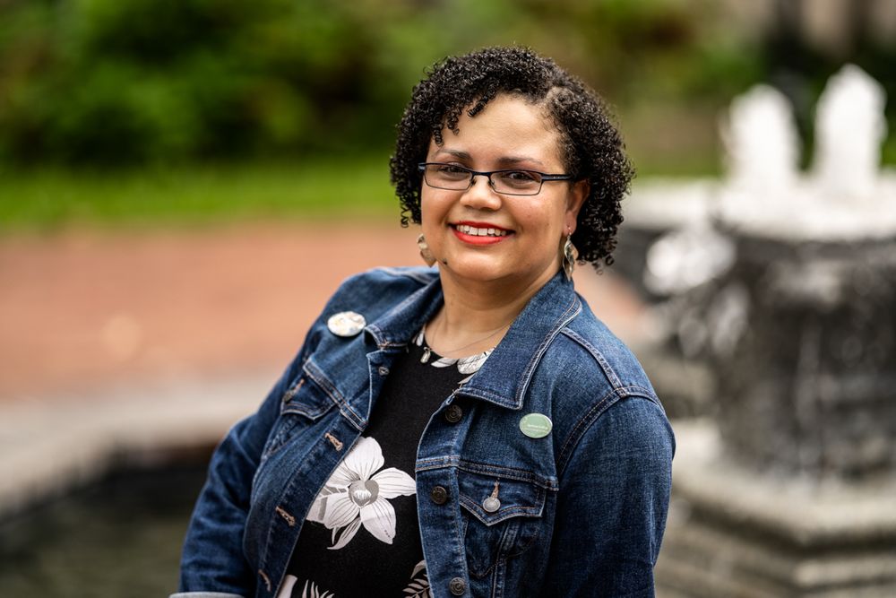 A photo of Kaia Alderson sitting, smiling in front of a fountain. Kaia is an African American woman. In the photo she wears glasses, a black and white flowery dress, a blue denim jacket, and dangly earrings.
